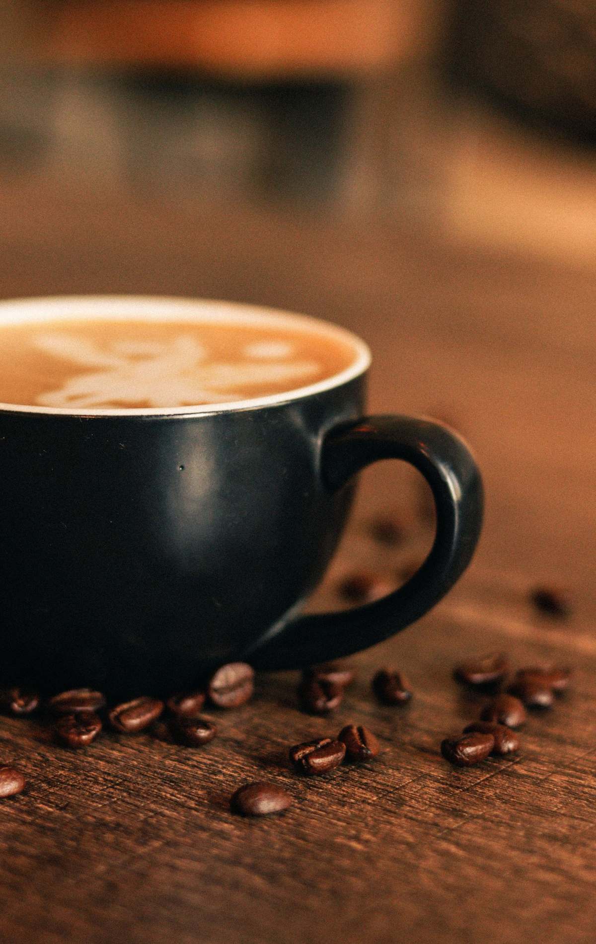 A latte with latte art in a dark cup, with coffee beans scattered on a wooden table