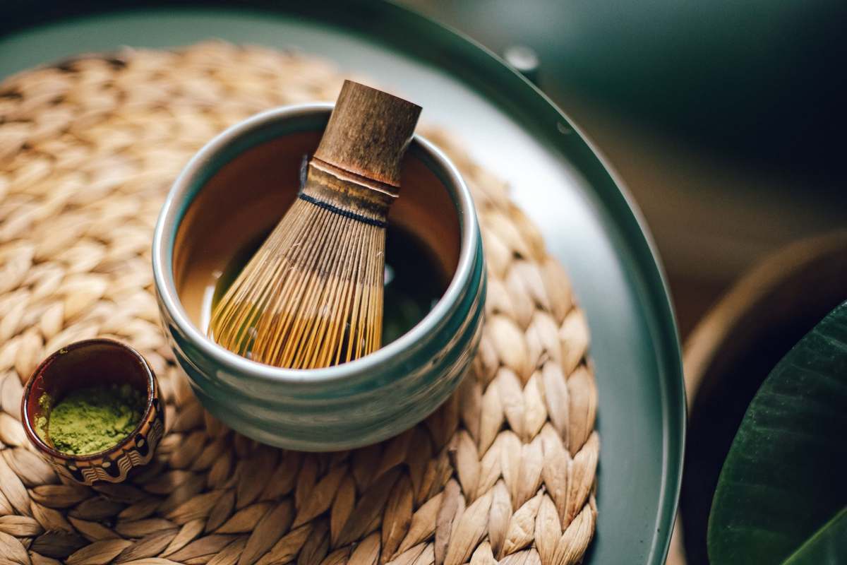 A bamboo matcha whisk in a ceramic bowl, with a small cup of matcha powder beside it