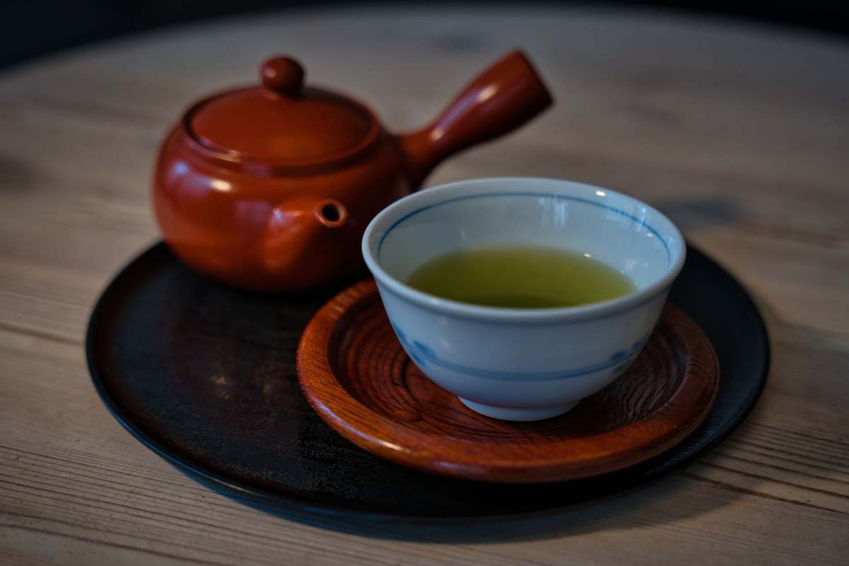 A cup of green tea on a wooden saucer beside a clay teapot on a wooden table
