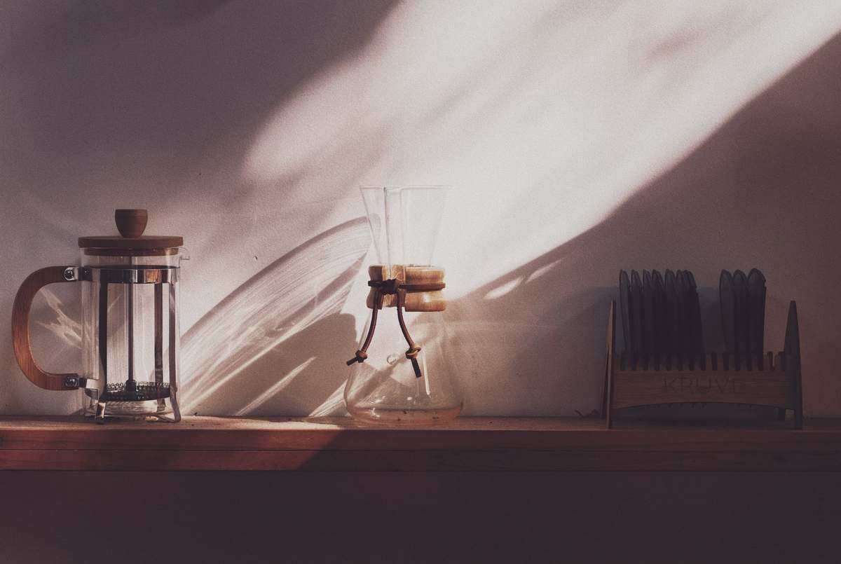 A cafetière and a pour-over coffee maker on a wooden shelf in soft window light